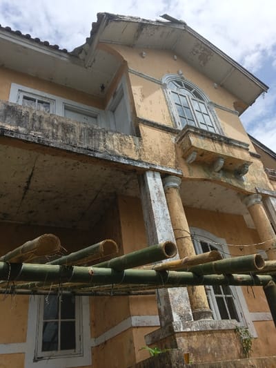 An upward-angled shot reveals a dilapidated, two-story structure located in Bình Hải, Vietnam, characterized by faded ochre-colored stucco and extensive signs of disrepair. The building features a prominent first-floor balcony with a concrete railing, supported by two heavily stained cylindrical columns. Above the balcony, on the upper level, an arched window with white multi-pane framing is visible on the right, accompanied by a smaller, decorative balustrade with corbels below it. To the left of the arched window, a rectangular window and a doorway with white frames open onto the main balcony. The roof, partially visible, exhibits terra cotta tiles and deteriorating white-painted eaves with peeling paint and structural damage. The building's surfaces display widespread paint peeling, dark biological staining consistent with mold or mildew, and general material degradation. In the immediate foreground, a trellis constructed from several green and dried bamboo poles, lashed together with dark wire or twine, partially obstructs the lower portion of the building. The sky above is overcast with scattered white clouds.