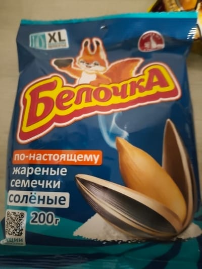The image shows a close-up of a bag of sunflower seeds. The bag is blue with red and yellow lettering. The brand name appears to be "Белочка" (Belochka). There is an image of a squirrel on the bag, and the words "по-настоящему жареные семечки солёные" (truly roasted salted seeds) are visible. The weight of the product is indicated as "200г" (200g). A QR code is also present on the bag. The bag is lying on a surface that appears to be a table or counter. There is no indication of the city of Васильево, Russia, in the image itself.