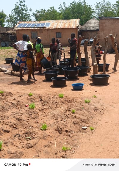 In Gidan Kogi, Nigeria, a group of people, including women and children, gather around a well and a collection of black plastic tubs. They appear to be drawing water from the well and filling the tubs. The scene takes place in a dirt-covered area with sparse vegetation, and a mix of traditional mud brick and corrugated metal roofed buildings, one with solar panels, are present in the background.