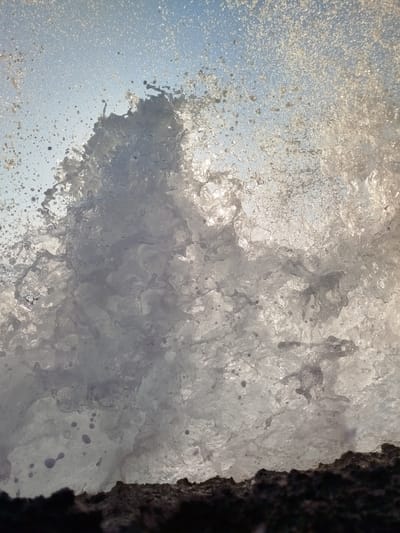 An extreme close-up shot captures a wave cresting and crashing against a dark, rocky shore. The water is rendered in a dynamic explosion of white foam and droplets, backlit by the bright sky, creating a translucent and ethereal effect. Small, rounded droplets of water are suspended mid-air. The sky above is a pale blue, speckled with even finer water mist. The dark, textured foreground suggests the jagged edge of rocks or a cliff face. The overall impression is one of raw, natural power and movement. The location is specified as Karaburun, Turkey, implying a coastal environment. No people are visible in the image.
