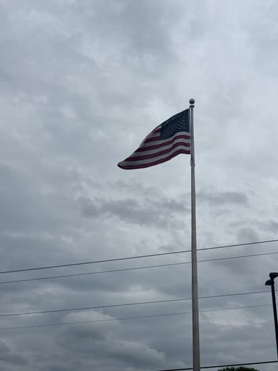 The image shows an American flag attached to a flagpole, flying against a cloudy sky. Power lines run vertically across the right side of the frame. Some greenery is visible in the upper right corner. The location is the city of Stone Mountain, United States of America.