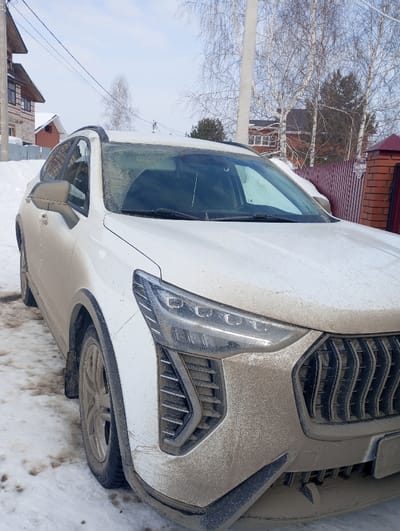 A white Haval SUV, heavily coated in brown road grime and mud, is parked outdoors on a snow-covered and muddy surface. The image captures the front-right quarter of the vehicle, showing its modern LED headlight, large grille, and a portion of the windshield. The foreground features dirty snow and exposed ground. In the background, a residential street scene unfolds with a two-story house on the left, featuring brick and light-colored siding. A wooden utility pole with power lines runs overhead. To the right, a reddish-brown metal fence with a brick post and another house with a dark roof are visible. Bare deciduous trees and some evergreen trees populate the background under a clear, light blue sky. The scene is located in the city of Яромаска, Russia. No individuals are present.