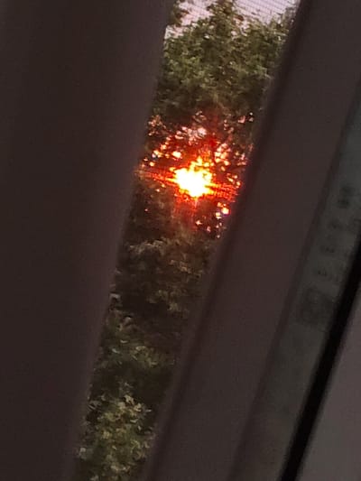 The image depicts a close-up view of a sun flare through foliage, with a dark, out-of-focus foreground and background that suggests an indoor environment, possibly a window. The sun is a bright, intense orange-yellow circle with visible rays, partially obscured by green leaves. The lighting suggests it is either sunrise or sunset. The image is grainy and slightly blurred, giving it an abstract quality. There is no discernible location in the city of Okhtyrka, Ukraine, or any people, subjects, or specific objects that would allow for that identification. The overall scene is ambiguous and lacks concrete details beyond the visual phenomena of light and shadow.