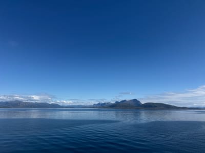 The image depicts a vast expanse of deep blue water under a clear, bright blue sky. In the distance, a rugged mountain range with some snow-capped peaks stretches across the horizon. The mountains appear to be covered in green vegetation in their lower slopes, and their peaks are sharp and imposing. The water is calm, with gentle ripples reflecting the colors of the sky and the distant landscape. The overall impression is one of serene natural beauty. This scene is likely located in the vicinity of Lødingen, Norway, a municipality known for its stunning coastal scenery and dramatic mountainous terrain.
