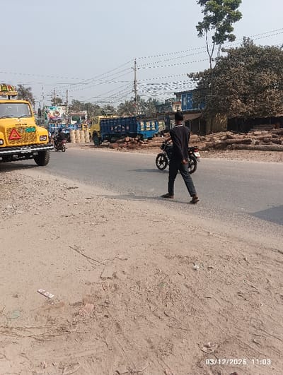 The image displays an outdoor scene in Kaliganj, Bangladesh, at 11:03 on March 12, 2025. The foreground consists of dry, light brown soil with scattered small debris. A paved asphalt road occupies the midground. On the left side, a yellow heavy-duty truck, equipped with a red triangular hazard sign on its grille, is partially visible on the dirt shoulder next to the road. A male subject, dressed in a dark long-sleeved shirt and dark trousers, is captured walking from left to right across the paved road, positioned beside a dark motorcycle. Another individual on a motorcycle is observed further down the road on the left. In the mid-distance, two blue cargo trucks are present on or near the road. A large pile of reddish-brown earth or construction material is situated between the road and the background structures. To the right, a significant stack of cut logs rests on the ground, adjacent to a large, leafy tree. The background features multiple utility poles supporting a dense network of overhead power lines against a clear, light blue sky. Indistinct buildings and additional trees are visible along the distant horizon.