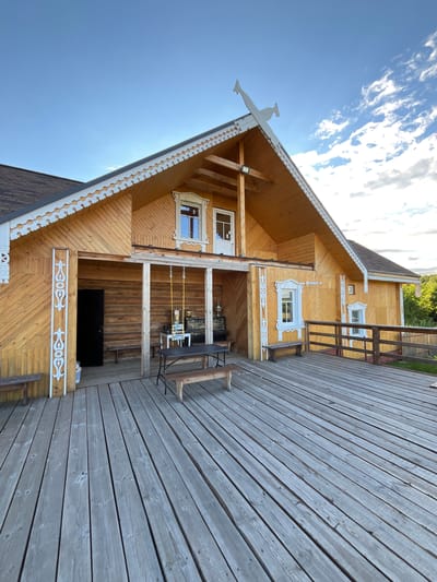 The image shows a wooden house with a porch in Vybolovo, Russia. The house is made of light-colored wood with ornate white trim around the windows and eaves. The porch is also made of wood and has a simple wooden railing. There are no people visible in the image.

The scene appears to be set during the daytime, with a bright blue sky and some clouds. The sun is shining, casting shadows on the house and porch. The overall impression is one of a rustic, peaceful setting.

Notable details include the intricate white carvings on the house, which suggest a traditional Russian architectural style. The wooden planks of the porch are weathered, indicating that they have been exposed to the elements. There are some chairs and a table on the porch, suggesting that it is used for outdoor relaxation. A black flat-screen television is visible under the porch. In the background, trees and greenery can be seen, indicating a rural or semi-rural location.