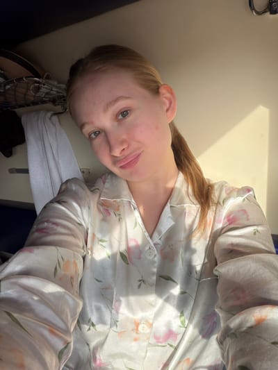 A fair-skinned person with reddish-blonde hair is lying down, wearing a light-colored, floral-patterned, button-down pajama top. The background appears to be a light-colored surface, possibly a wall or ceiling, with a hint of natural light. A gray towel hangs off a metal frame. The location in the city of Krasnyy Sulin, Russia is not determinable from the image.
