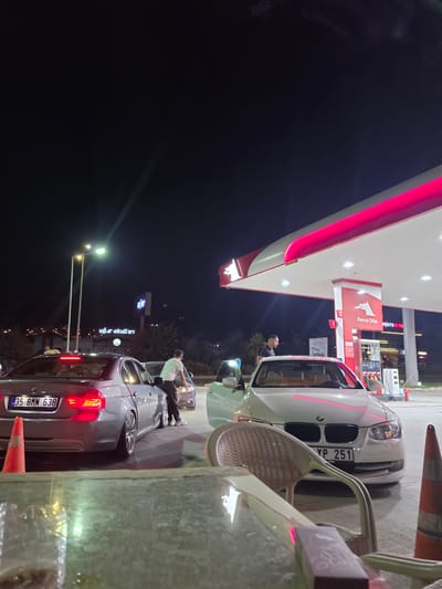 The image depicts a night scene at a gas station in Güzelbahçe, Turkey. In the foreground, a table with a reflective surface and chairs are visible, suggesting an outdoor seating area. Further back, a grey BMW sedan with license plate "35 BMN 638" is parked, its taillights illuminated. To its right, a white BMW sedan is positioned at a gas pump. A man in a white t-shirt and dark pants is standing between the two cars, appearing to interact with the white BMW. The gas station features a red and white canopy illuminated with pink LED lights, with "Petrol Ofisi" signage visible on a pump. The background shows the dark night sky, distant city lights, and trees. The overall atmosphere is that of a typical roadside establishment operating after dark.