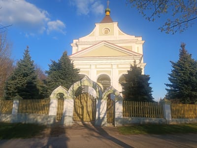 The image presents a sunlit view of an Orthodox church or chapel, situated in Шчорсаўскі сельскі Савет, Belarus. The main subject is the church, a white and light yellow building featuring classical architectural elements such as columns, arches, and a triangular pediment. A small, green-domed cupola topped with an Orthodox cross sits atop the roof. On the upper facade, within a circular frame, there is an icon or fresco depicting a bearded face, likely Jesus Christ or a saint.

The church is flanked by tall, dark green evergreen trees on both sides. In the foreground, a light-colored wooden picket fence with a low concrete foundation encloses the church grounds. Two arched gate structures, matching the light green and white color scheme of parts of the church, form the entrance, with wooden gates. A paved path or road runs in front of the fence, with green grass visible on the sides.

The scene is captured during the golden hour, either early morning or late afternoon, indicated by the long shadows cast across the path and fence by the fence posts and other objects. The weather is clear and sunny, with a vibrant blue sky featuring a few scattered white clouds. Bare branches of deciduous trees, some with small green leaves, are visible in the upper parts of the frame, suggesting springtime. No people or specific activities are depicted, contributing to a tranquil and serene atmosphere in this rural or small-town setting.