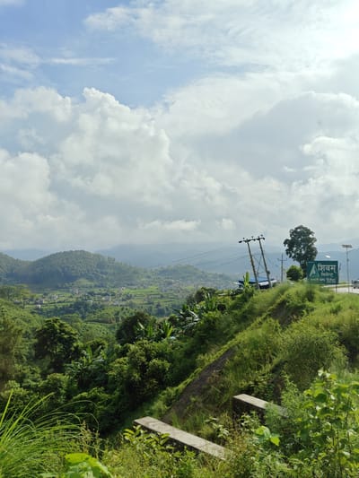 The image depicts a lush, green hillside in Tharpu, Nepal, with a panoramic view of a valley and distant mountains under a cloudy sky. The foreground is dominated by dense vegetation, including tall grasses and leafy bushes, with two concrete ledges visible. A dirt path runs along the hillside. In the middle ground, a village is nestled amidst terraced fields, and a single large tree stands prominently. Power lines stretch across the scene, supported by utility poles. A green sign with text in Devanagari script, reading "शिवम् सिमेन्ट" (Shivam Cement), is mounted on a pole. A white vehicle is partially visible on a road in the background. The overall impression is of a serene, rural landscape.