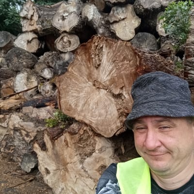 A man wearing a dark patterned bucket hat and a neon yellow safety vest smiles at the camera, positioned in the lower right of the frame. He is in front of a large pile of chopped wood, with trees visible in the background. The man's face is the primary subject, with the wood pile serving as the backdrop. The scene appears to be outdoors, possibly in a rural or forested area. No specific location within Montgailhard, France, is discernible from the image.
