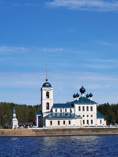 The image captures a Russian Orthodox church complex situated on the banks of a body of water, possibly a river or lake. The church is located in Koprino, Russia. The complex is composed of several interconnected structures, including a main church building with multiple blue onion domes, a bell tower, and lower auxiliary buildings. The buildings are predominantly white or light yellow with green roofs. A short wall runs along the water's edge, separating the complex from the water. The background consists of a dense forest. The sky is a clear blue with scattered clouds. The water in the foreground is dark blue. A monument, possibly a memorial or marker is to the left. An army truck is parked near the monument.