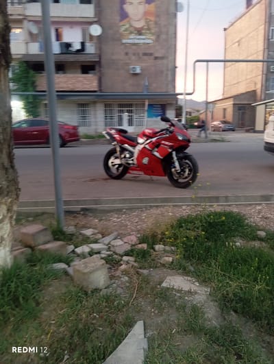 This is a vertically oriented, slightly blurry photograph taken in Vedi, Armenia. The main subject is a vibrant red motorcycle parked on a paved surface, likely a street or sidewalk. The motorcycle is positioned facing away from the camera, with its front wheel turned slightly to the left. It appears to be a sportbike with sleek lines and bold graphics, including some white lettering visible on its side.

To the left of the motorcycle, a red car is partially visible, suggesting it is parked nearby. Behind the motorcycle and car, there is an apartment building with multiple windows and balconies, indicative of an urban residential area. A large poster or advertisement featuring a person's face is mounted on the side of the building. A satellite dish is also visible on the building's facade.

The scene is bordered by a strip of grass and some scattered rocks and debris on the right side. The lighting suggests it might be late afternoon or early evening, with soft, diffused light and a pale sky. The overall impression is a snapshot of daily life in a residential neighborhood. The text "REDMI 12" is visible in the bottom right corner of the image, indicating the device used for capturing the photo.