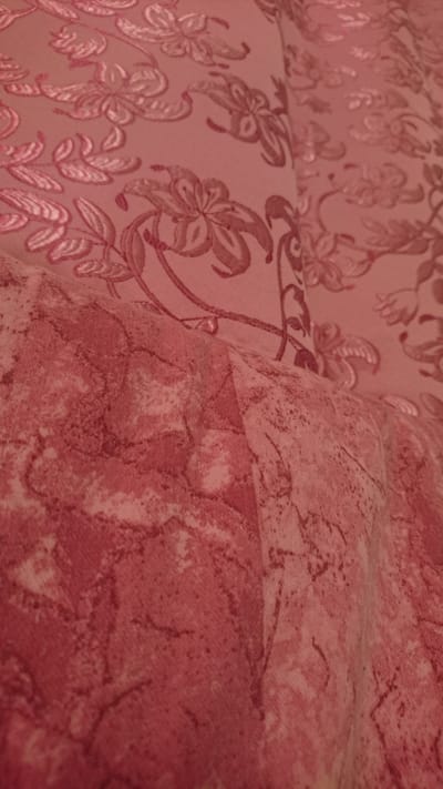 The image shows a close-up view of two pieces of pink fabric with floral patterns. The fabric in the upper portion of the image has a lighter pink background with darker pink floral and leaf designs. The fabric in the lower portion has a more mottled appearance, with shades of pink and reddish-brown, resembling a marble or tie-dye effect. The fabrics appear to be draped or folded, creating shadows and highlights that emphasize the textures and patterns. There is no indication of any location in the city of Dvirets, Ukraine, or any people, actions, or interactions depicted in the image.