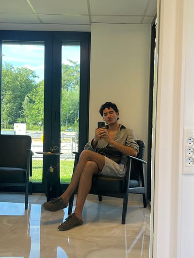 A young man with dark, curly hair is seated in a black armchair, holding a smartphone. He is wearing a striped button-down shirt, open at the collar, and light-colored shorts. His legs are crossed, and he is looking at the phone, with one eye winking. The armchair is positioned in front of a large window that looks out onto a green, grassy area with trees and some buildings. To the left of the armchair, another black armchair is partially visible. In front of the man, on a small table shaped like a cat with glasses, are a pair of glasses and a small object. The floor is tiled with a polished marble pattern. To the right of the man, a white wall features a power outlet with three circular sockets. The scene appears to be an indoor waiting area or lounge, possibly at a business or public facility in Plainevaux, Belgium, given the window view of a semi-rural or suburban landscape.