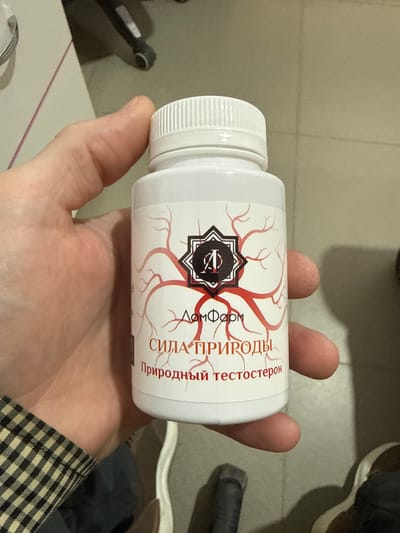 A white plastic bottle with a white screw-on cap is held in a person's hand. The bottle has a label with red and black text and graphics. The text on the label appears to be in Russian and includes the words "ДОМФАРМ", "СИЛА ПРИРОДЫ", and "Природный тестостерон". The graphics on the label depict a stylized red vascular network. The background of the image shows a tiled floor and a light-colored wall. The image does not contain any information indicating a location in Mayrtup, Russia.