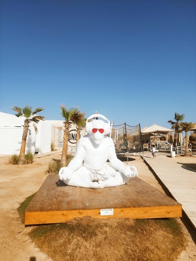 The image depicts a sunny outdoor scene at a beach resort in Ras Abu Soma, Egypt. In the foreground, a white, life-sized statue of a monkey in a meditative pose, wearing red sunglasses and white headphones, sits on a wooden platform. The statue is positioned near a white building with the letters "UNI" visible. In the background, there is a trampoline with people on it, along with tables, chairs, umbrellas, and palm trees, suggesting a beachside recreation area. A wooden walkway runs along the right side of the image. The sky is clear and blue. A small sign can be seen attached to the front of the wooden platform on which the statue sits. There is a child running in the background.