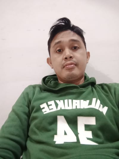 A medium-shot, low-angle portrait of a man wearing a green hooded sweatshirt with the white text "MILWAUKEE" and "AE" printed on it. The man has dark hair styled with a slight wave on top, light brown skin, and a light mustache and goatee. The background is a plain, off-white wall. The location is Cangkring, Indonesia.