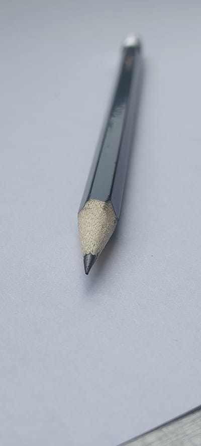 A sharpened graphite pencil with a hexagonal gray barrel rests diagonally on a white, textured surface, likely paper. The pencil's tip is pointed and dark, with visible graphite. The wood casing around the graphite is exposed and appears light brown. The background is a plain white, with a slight shadow cast by the pencil. There are no people or other objects visible in the frame. The image is a close-up, with shallow depth of field, blurring the background. The scene does not depict any recognizable urban or natural landscape elements, and therefore, its location within Storozhevaya-2, Russia, or any other city, cannot be determined from the visual information provided.