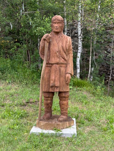 A full-body wooden sculpture of a man stands upright on a grassy area in Srednie Kichi, Russia. The sculpture depicts a man with a weathered face, dressed in what appears to be traditional clothing, including a tunic and boots. He is holding a long wooden staff in his right hand, resting it on his shoulder. The sculpture is positioned on a concrete base which is surrounded by green grass. In the background, a dense forest of trees, including birch trees with white bark, rises up a rocky incline. The sky is overcast.