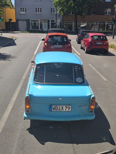 The image shows a street scene in Wittenberg, Germany. A light blue Trabant 601 is parked on the right side of the road. Next to it is a red Honda Jazz. Further ahead is a red Ford Fiesta. Buildings line the left side of the street. A traffic light is visible in the distance. The focus is on the rear of the Trabant, highlighting its "DDR" sticker and license plate "WB X 79." The road surface is asphalt.