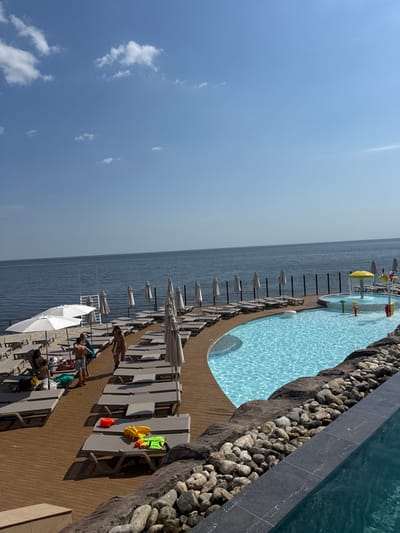 The image depicts an outdoor resort area, identified by the prompt as located in Hlibivka, Ukraine. The scene is illuminated by daylight under a clear blue sky with sparse white cumulus clouds. A large, calm body of water, consistent with a sea or large lake, forms the horizon line and extends across the background.

In the mid-ground, a light-brown wooden deck functions as a sunbathing area. Numerous grey, padded lounge chairs are arranged in rows on this deck, with several beige, folded beach umbrellas positioned between them. To the far left, two large white open umbrellas are providing shade over occupied lounge chairs. A dark, vertical slat fence separates the wooden deck from the large body of water.

Adjacent to the wooden deck is a large, irregularly shaped swimming pool filled with clear, blue water. Sunlight reflects off the water's surface, creating ripple patterns. Connected to this main pool, nearer the water's edge, is a smaller, circular children's pool featuring a yellow mushroom-shaped water sprayer and several colorful, smaller water spouts. In the foreground, the edge of another pool with dark grey tiling is partially visible on the bottom right. A rugged rock formation, interspersed with lighter river stones and pebbles, forms a landscaped border between the foreground pool and the wooden deck/main pool.

Approximately eight individuals are present on the wooden deck and in the main pool. Most are adults or young adults, dressed in swimwear. A group of four to five individuals is gathered on the left side of the deck, some standing, some seated on loungers. One individual in a light-colored swimsuit is walking across the deck towards the right. Another person is partially submerged in the main swimming pool. On a set of unoccupied lounge chairs in the lower-center, brightly colored plastic water toys, including an orange bucket, a yellow shovel, and green water guns, are visible. The scene appears to be an active recreational environment.