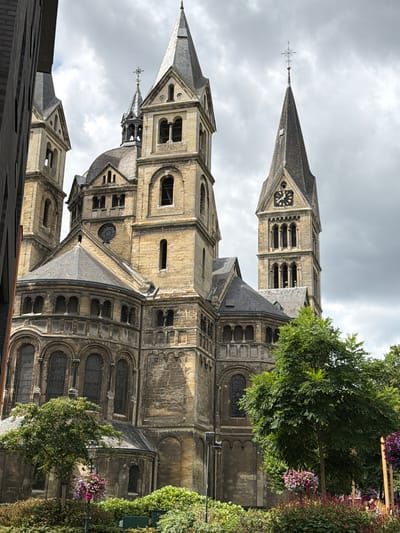 The image shows the exterior of the Saint Christopher Cathedral in Roermond, Netherlands. It is a large, imposing stone structure with multiple spires and towers. The architecture is Romanesque and Gothic in style, with arched windows and intricate stone carvings. The cathedral is surrounded by lush green trees and flowering bushes. The sky is overcast, suggesting a cloudy day. There are no people visible in the image. The perspective is from a low angle, looking up at the cathedral.