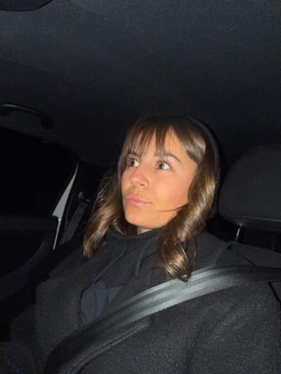 A young woman with bangs and brown hair sits in a car, wearing a seatbelt. She looks towards the camera with a slight pout. The interior of the car is dark, with only the woman's face and part of the seat visible. The lighting suggests it's nighttime.