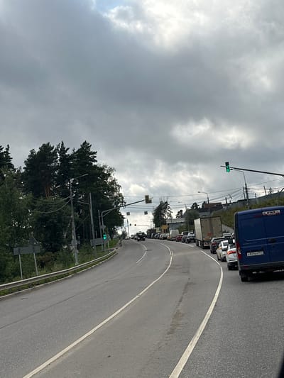 The image depicts a traffic jam on a cloudy day. The scene is on a multi-lane road with a barrier on the left side. Several vehicles, including trucks and cars, are visible in the queue. Trees and power lines line the side of the road. A traffic light with a green signal is visible in the upper right portion of the image. The location is identified as Aristovo, Russia. The overall atmosphere is one of congestion and subdued natural light.