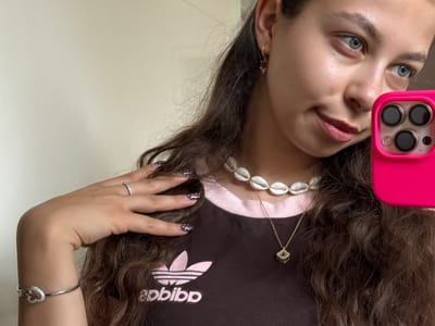 A young woman with long curly brown hair is taking a selfie. She is wearing a brown t-shirt with a pink Adidas logo, a seashell choker necklace, and a gold pendant necklace. She is also wearing earrings, a ring, and a heart-shaped bracelet. Her nails are painted with a white polka-dot design. The phone she is using to take the selfie is pink. The background is a plain, light-colored wall. There is no indication that this image was taken in Bruchköbel, Germany.