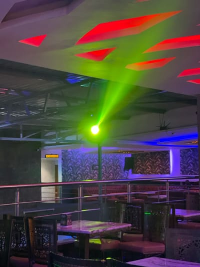 This media file captures the vibrant interior of what appears to be a modern restaurant, bar, or lounge, likely at night, in Kimorori, Kenya. The scene is dominated by striking artificial lighting.
On the left side of the frame, a light-colored wall features bold, angular cutouts that are illuminated from behind with a bright red glow, creating a geometric pattern. A powerful green spotlight is prominent in the mid-left, casting a focused beam of intense green light across the space.
To the right, the establishment is furnished with several tables and chairs. The chairs have light-colored, possibly upholstered, seats and backs, with intricate dark metalwork patterns forming their frames and supports. The tabletops appear to be made of a light-colored material, possibly marble or stone. Behind the seating arrangement, walls with textured or patterned finishes are visible, softly lit with blue and purple hues, enhancing the overall atmospheric lighting. A metallic railing structure, possibly a staircase or a decorative partition, runs vertically through the right side of the image.
There are no people visible in the image, and no specific activity is taking place beyond the general setting of a dining or social venue. The combination of intense, colored spotlights and ambient lighting suggests a lively and contemporary atmosphere. In the mid-ground, on a wall, an indistinct yellow-orange sign or text is visible, but the characters are too pixelated to be legible.