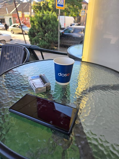 A digital photograph captures a close-up view of a glass table outside a cafe, with a blurred street scene in the background. On the table are a smartphone with a dark screen, a pack of Parliament Aqua Blue cigarettes with a lighter resting on top, and a blue disposable cup with the white logo "dose" printed on it. The background shows a street with passing cars and pedestrians, and trees. The scene suggests a relaxed outdoor setting in Khasavyurt, Russia, where someone is taking a break.