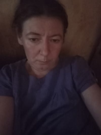 The image shows a woman from the chest up, looking down. She has dark hair and is wearing a blue shirt. The background is a blurred, textured surface, possibly a wall or furniture. The lighting is dim, creating shadows on her face. The location in Berezivka, Ukraine, is not identifiable from the image itself.