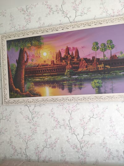 The image shows a framed painting of a temple complex, possibly Angkor Wat, in Cambodia. The painting depicts the temple at sunset, with a vibrant sky of orange, pink, and purple hues. The temple is reflected in the calm water in front of it, and a large tree with hanging vines is visible on the left side of the painting. Palm trees are also present on the right. The painting is hung on a wall covered with floral wallpaper, featuring a pattern of light pink cherry blossoms on a white and grey textured background. The frame of the painting is ornate and cream-colored with decorative detailing. The overall impression is one of a decorative interior scene. There are no people or animals visible in the image. The location is not specified as Афонино, Russia.