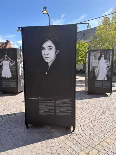 The image depicts an outdoor exhibition of large-format black and white photographs displayed on black panels. The exhibition is set in a paved public space, possibly a square or plaza, in Lund, Sweden. The ground is paved with cobblestones arranged in a concentric pattern. In the background, there are buildings with red tile roofs, trees with green foliage, and a bright blue sky with wispy clouds. Streetlights are visible, mounted on poles extending over the exhibition panels.
The central panel features a portrait of a woman with dark hair and expressive eyes, looking directly at the camera. Text is printed below the portrait, likely providing biographical information and context for the photograph. To the left and right of the central panel, other exhibition panels are visible. The panel on the left shows a woman with long hair, wearing a white dress. The panel on the right shows a woman draped in a white cloth. The overall impression is one of an art installation in a public urban setting.