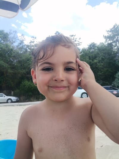 A young child, with dark hair and brown eyes, is pictured on a sandy beach. The child is facing the camera with a slight smile and has their right hand touching their forehead. They are wearing an earring in their left ear. Sand is visible on their bare chest and shoulders.

In the background, out-of-focus trees and a blue sky with clouds are visible. A beach umbrella with blue and white stripes is partially visible in the upper left corner. Cars are parked in the background, suggesting a public beach. The presence of sand and the child's attire suggest a daytime beach outing. The overall mood is cheerful and relaxed, indicative of a pleasant day. The location is Osenovo, Bulgaria.