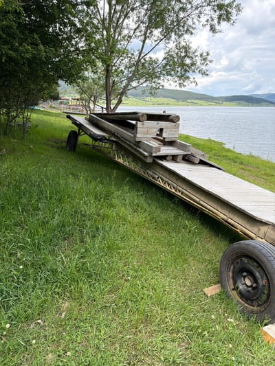 This image, captured in Tsigov chark, Bulgaria, presents an outdoor scene featuring a large, inclined wooden structure positioned on a grassy bank next to a lake. The wooden structure, likely a disassembled pier or boat ramp, is elevated on a metal frame and supported by two visible black tires with rusty rims; one tire is prominent in the bottom right, resting on a wooden block, while another is partially obscured in the mid-left. Various wooden planks and logs are stacked unevenly on top of the main structure. A single individual with dark hair and a dark top is partially visible behind these stacked timbers, looking towards the left side of the lake.
The foreground consists of lush green grass with scattered small white flowers. To the left, dense green foliage from trees and bushes obscures the background. A large tree with a prominent trunk and sparse green leaves stands in the mid-ground, slightly right of center. The smooth surface of a lake occupies the middle and right sections of the image. Across the lake, rolling green hills, partially covered with trees, extend into the distance under a cloudy sky with varying shades of grey and white. In the far distance on the left side of the lake, faintly visible through the trees, are several structures with colorful roofs and a small pier.