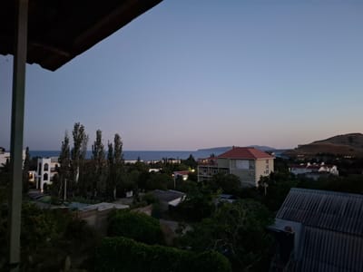 The image depicts a twilight view of a coastal settlement, likely in Щебетовский поселковый совет, Ukraine. The sky is a gradient of pale blue to a lighter hue near the horizon, suggesting the sun has recently set. In the foreground, a dark, angled structure, possibly a roof or balcony overhang, partially obscures the upper left portion of the frame. The middle ground is filled with a dense arrangement of trees, varying in height and leafiness, interspersed with buildings. These buildings appear to be residential, with some featuring multiple stories, balconies, and pitched roofs. A body of water, likely the sea, is visible in the distance, stretching across the horizon. Beyond the settlement and water, a gently sloping hillside with sparse vegetation and a few distant lights can be seen on the right. The overall atmosphere is tranquil and serene, characteristic of early evening in a rural or semi-rural coastal area. There are no people visible in the image.