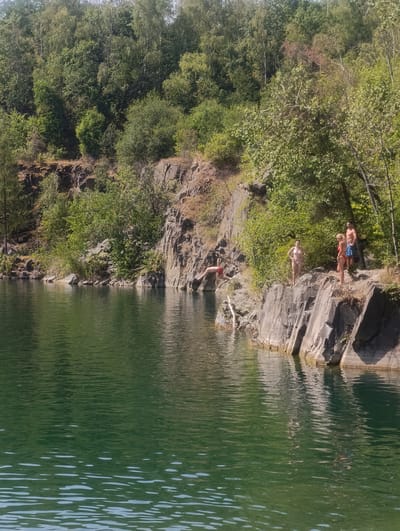 The image captures a scenic quarry in Výkleky, Czechia, on a sunny day. The quarry features a deep, emerald-green body of water surrounded by steep, rocky cliffs covered in lush vegetation. On the right side of the quarry, a group of four people are present. One person, a male wearing red shorts, is in mid-air, having just jumped off the cliff into the water. Three other individuals, two females in bikinis and one male wearing blue swim trunks, stand on the rocky edge, observing the jump. The water is calm, reflecting the surrounding greenery and rocky formations. The overall scene depicts a recreational activity in a natural environment.