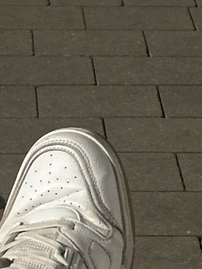 The image shows a close-up view of the toe area of a white sneaker resting on a textured, dark grey surface. The surface appears to be made of rectangular paving stones or tiles, arranged in a grid pattern with visible grout lines. The sneaker is white with perforations on the toe box, characteristic of athletic footwear. Stitching details are visible along the seams of the shoe, and the sole has a raised tread pattern. The lighting suggests it is daytime, with shadows casting from the shoe onto the paving stones, indicating a sunny or brightly lit environment. The perspective is from a high angle, looking down at the shoe and the ground. There is no visible text in the image. The setting appears to be outdoors, possibly a street, sidewalk, or patio in Siedlce, Poland. No specific activity or event is depicted, but the image focuses on the shoe and its immediate surroundings, suggesting a moment of pause or a detail shot.