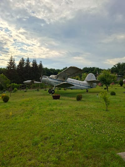 The image depicts an outdoor scene in Malcoci, Moldova, featuring a vintage Antonov An-2 biplane parked on a grassy lawn. The plane is painted light blue and white, with visible markings that include "CCCP 32424". It is positioned on its landing gear. The surrounding environment is a well-maintained park or garden with lush green grass dotted with small white flowers. Several trees, including evergreen conifers and deciduous trees, are visible in the background, along with a few bushes and small ornamental trees in the foreground. In the distance, part of a building with a red brick chimney can be seen, suggesting the presence of structures nearby. The sky is overcast with a mix of light and dark clouds. The overall impression is that of an open-air display or a historical artifact preserved in a tranquil setting.