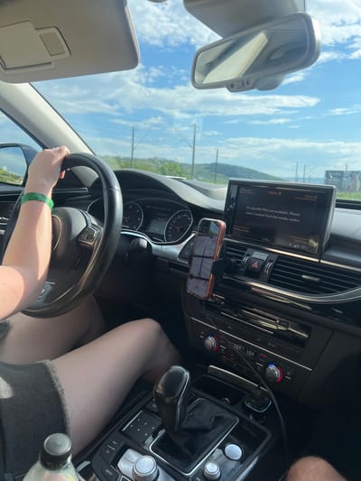 An exterior shot from inside a vehicle shows a person's hands on the steering wheel. The hands are positioned at the 10 and 2 o'clock positions on a black steering wheel. The person is wearing a green bracelet on their left wrist. The interior of the car is visible, including the dashboard with a central infotainment screen displaying a navigation interface and climate controls. The screen shows a message "No playable files are available. Please select content from the media centre." A smartphone mounted on a holder on the dashboard displays a navigation app. The climate control panel shows a temperature of 18.0 degrees Celsius. Outside the car, a bright, cloudy sky is visible, with hints of greenery and distant hills suggesting a rural or semi-rural environment. The image is taken from the passenger seat looking towards the driver's side. The scene is consistent with a person driving a car on a road in the vicinity of Sânnicoară, Romania, given the visible landscape and typical vehicle interior.
