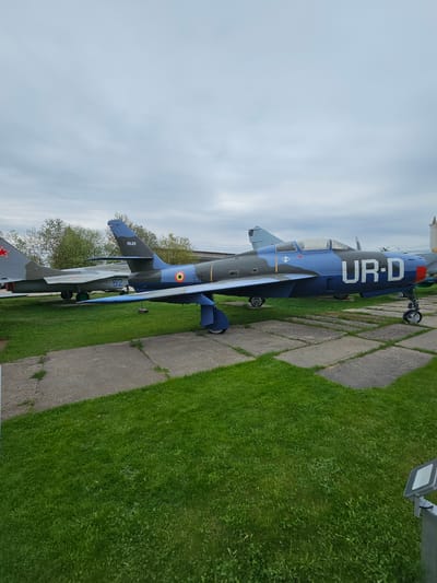 The image shows two military aircraft on display at what appears to be an outdoor museum or park. The location is most likely in the city of Arkhangel’skoye, Russia, given the context provided.

The primary aircraft in view is a blue and gray jet with the letters "UR-D" in white on its side. It has a red nose. It's positioned on a concrete slab alongside a patch of green grass.

Behind it, a second aircraft, painted in gray and green camouflage, is partially visible. It has a red star on its tail section, suggesting a Soviet or Russian origin.

The aircraft are stationary, presumably part of a static display. The background includes trees and a cloudy sky. A light fixture is visible on the right side of the image. The ground consists of grass and a concrete path.