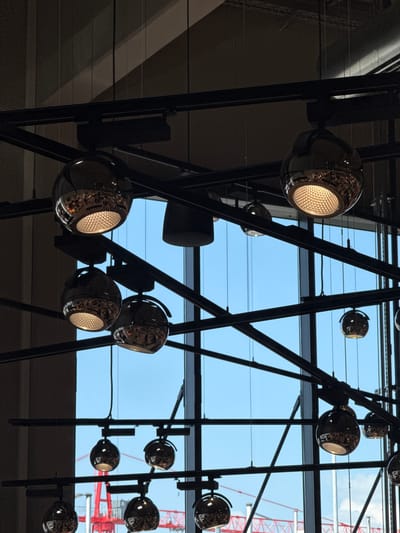 This image captures a cluster of modern pendant lights hanging from a dark, angular metal structure against a backdrop of a bright blue sky. The lights themselves are spherical, with a metallic, reflective surface that mirrors the surroundings. Some of the lights are illuminated, casting a warm, yellowish glow and revealing intricate patterns within their reflective casings. Others are unlit, appearing as dark, polished spheres. The metal structure is composed of thin, dark rods and beams, creating a geometric, almost architectural framework.

Beyond the structure and lights, the bright blue sky suggests it is daytime, likely clear with minimal cloud cover. The presence of a red construction crane in the upper right quadrant of the image provides a strong cue to the location, indicating an area undergoing development or construction. The overall scene evokes a sense of modern design and industrial aesthetics, possibly within a large public space, atrium, or exhibition hall. There are no people visible in the image, and no text is apparent. The angle of the shot is from below, looking upwards towards the ceiling and the sky beyond.