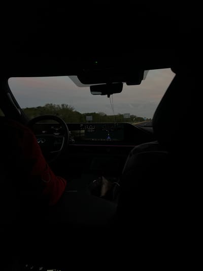 The image presents the interior perspective from the rear passenger seat of a vehicle in motion, looking forward through the front windshield onto a highway scene in Pomuch, Mexico. The ambient light outside suggests evening or dusk, with an overcast sky.
Inside the vehicle, the primary illumination originates from a wide, dual-screen digital dashboard display. The steering wheel, featuring an Infiniti logo, is visible on the left. The driver's left arm, clad in a red long-sleeved garment, is positioned on the steering wheel. The driver's side display indicates "18:49" as the time and "157" (likely kilometers per hour) as the speed, along with a Spanish message "Aviso Actualmente no disponible" (Notice Currently not available). The larger central display, also showing "18:49," appears to be a navigation or infotainment system with various icons. A rearview mirror hangs from the ceiling, from which a string of beads or similar ornamentation dangles. A small green indicator light is visible on the overhead console. The dark silhouette of a front passenger's headrest and upper body is present on the right side of the frame. The windshield exhibits minor smudges or dust.
Outside the vehicle, a multi-lane highway stretches into the distance, flanked by dense green foliage and trees. Several distant highway signs are discernible. The sky above is a muted gray-blue, consistent with twilight conditions.