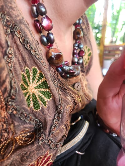 This image shows a close-up view of a person's neck and upper chest, adorned with a beaded necklace. The necklace features dark, iridescent pearls, interspersed with red beads and a large, polished tiger's eye stone. The person is wearing a brown, embroidered garment, possibly a tunic or dress, with intricate floral patterns in green and red thread. The embroidery is detailed, with a prominent green flower design in the center of the frame. The fabric appears to be a natural material, possibly linen or cotton, with a slightly textured weave. A black accessory, possibly a bag or clothing item with a zipper, is visible on the right side of the frame, adding a modern contrast to the ethnic-style garment. The background is softly blurred, showing hints of greenery and what might be natural light filtering through trees or foliage, suggesting an outdoor setting. The lighting indicates it is daytime, likely in the morning or afternoon. The overall mood is relaxed and casual, possibly during a leisurely outing in a warm climate like Hoi An, Vietnam, given the contextual information. There is no visible text in the image.