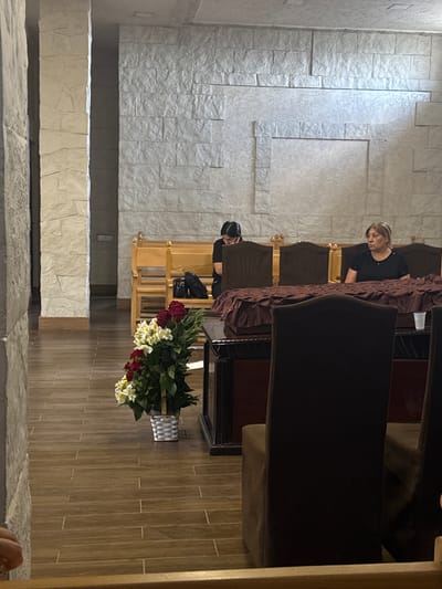 The image depicts the interior of a funeral home or chapel in Masis, Armenia. The room has walls covered in a textured, off-white stone-like material and a wooden floor. In the foreground, a large, dark-colored funeral casket, draped with a dark red cloth, is positioned prominently. Several dark brown chairs are visible around the casket, with one in the immediate foreground partially obscuring the view.

To the left of the casket, a basket of flowers, primarily red roses and white alstroemeria, is placed on the floor. In the background, there are wooden pews or benches. Two women are seated on these benches. The woman on the left is facing down, seemingly in prayer or contemplation, and has dark hair pulled back. The woman on the right, with blonde hair, is looking towards the right side of the frame. A black bag rests on the pew near the woman on the left. The overall atmosphere appears somber and respectful, consistent with a funeral service.