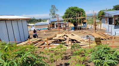 This outdoor image depicts an active construction or development site in Tchere, Democratic Republic of the Congo, under a bright blue, clear sky. In the immediate foreground, green, leafy plants, possibly cassava, grow in the reddish-brown soil, partially obscuring piles of wooden planks and building materials.

The mid-ground is dominated by several structures in various stages of completion. On the left, a building with white fabric or tarp walls and a corrugated metal roof appears largely finished. Nearby, three individuals are engaged in work: one person in dark clothing is bent over, seemingly arranging wooden planks, while another stands close by, and a smaller figure, possibly a child, is partially visible behind the bent worker.

Further into the scene, multiple other buildings are visible, characterized by blue corrugated metal roofs and light-colored walls. Many of these structures have solar panels installed on their roofs. Several wooden frameworks indicate ongoing construction. A large, green-leafed tree stands prominently in the mid-right. To the far right, another worker wearing a yellow safety vest is visible near a partially constructed building with white walls. White wooden fences or barriers are also present around parts of the site.

The overall scene portrays active community development or settlement expansion, featuring practical, adaptable architecture and an emphasis on renewable energy with the widespread use of solar panels. The distant background shows a hilly landscape with scattered trees. The image is watermarked with "Joseph Kisuki" in the bottom right corner.
