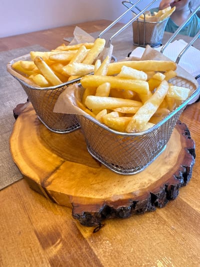 In a close-up, high-angle shot, two wire baskets filled with golden French fries are presented on a rustic wooden slab. The fries are uniformly cut and appear to be freshly fried, with some showing slight browning. The baskets are lined with brown paper, and one is slightly tilted, allowing some fries to spill over. The wooden slab has a natural, uneven edge with dark bark, showcasing the wood grain. In the background, a person wearing a light blue shirt is partially visible, with their hand reaching towards the fries. The lighting suggests an indoor setting, possibly a restaurant or dining area. The scene evokes a sense of casual dining and enjoyment.