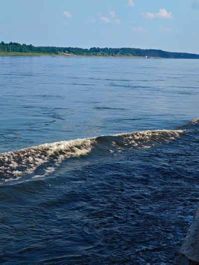 This is a high-angle, full shot of a wide, dark blue river. The river occupies the majority of the frame, with a foamy white wave in the mid-ground and a distant shoreline in the background. The shoreline is lined with a dense forest of green trees and a clear blue sky with a few scattered white clouds above. A small white boat is visible on the water near the distant shore. In the lower right corner of the image, a textured, greyish-brown object, possibly part of a dock or pier, is visible. The scene is devoid of people. The location is the city of Ust'-Kem', Russia.